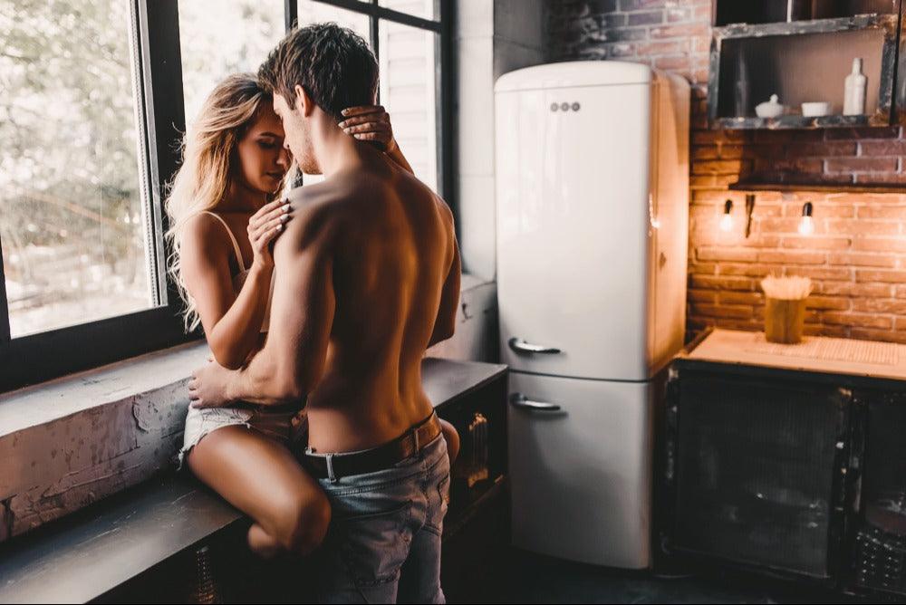 A women in shorts and tank top on counter with a man in jeans and no shirt holding her close 